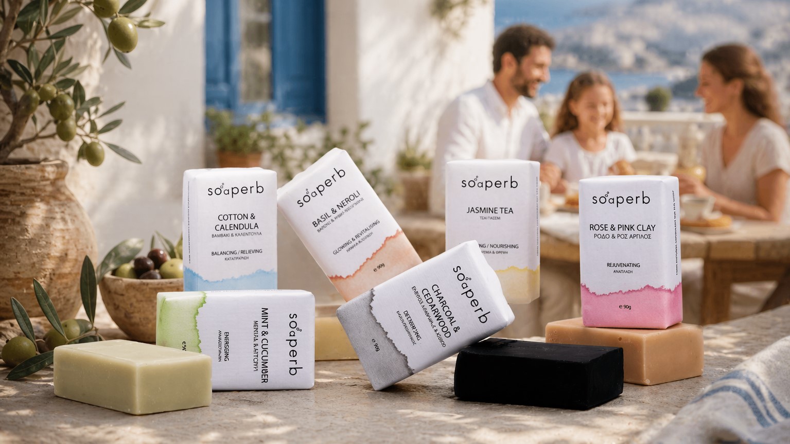 Přírodní řecká olivová mýdla Soaperb naaranžovaná na kamenném stole, doplněná o olivové větvičky a středomořské dekorace; v pozadí rozostřená rodina sedící u stolu v typickém řeckém prostředí s výhledem na moře.